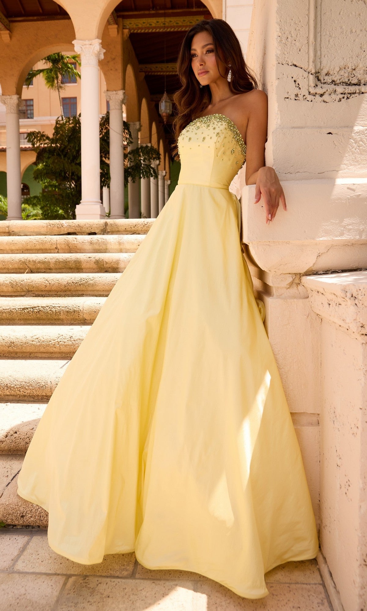 A woman in the Ava Presley 26417 long prom dress, featuring a strapless, floor-length yellow design with beaded detailing, stands by stone steps and columns outside a building.